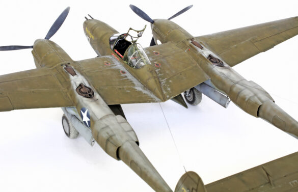 Photo Gallery – Tamiya 1/48 P-38G Lightning by Chris Wauchop Radio compartment detailing using 0.2 mm and 0.3 mm wire – painted placards and realistic wiring. Supercharger units finished with layered metallic and brown/black shades for realistic depth. Fuselage underside showing grime wash and added hydraulic brake lines. Drop tanks weathered for operational realism, mounted underwing. Panel line seal tape simulation using airbrush fading and masking techniques. Camouflage tones stippled for realistic weathered surface finish. Paint wear and black exhaust marks enhance visual realism. Cockpit close-up with foil-made harness and mirror wire control. Wing underside shows soot, staining, and sealing tape simulation. Tail surfaces display extensive weathering and exhaust wear effects.
